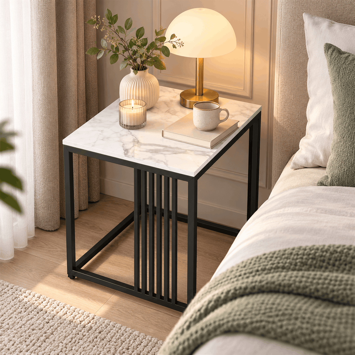Square white marble look side table with a black slatted frame beside a bed holding a lamp, a vase, a candle and a book