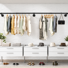 Long black industrial pipe wall mounted clothes rack with shirts and dresses on hangers and a handbag on a hook above drawers