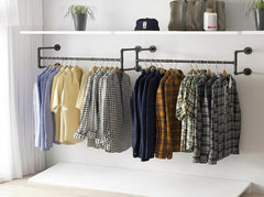 Two black industrial pipe wall mounted clothes racks side by side with plaid shirts on wooden hangers below a white shelf