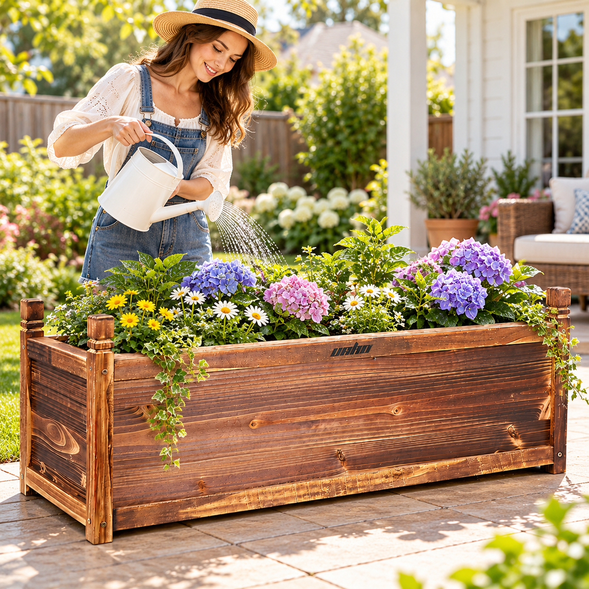 Woman in a straw hat watering flowers in a long wooden planter box with a white watering can on a sunny patio